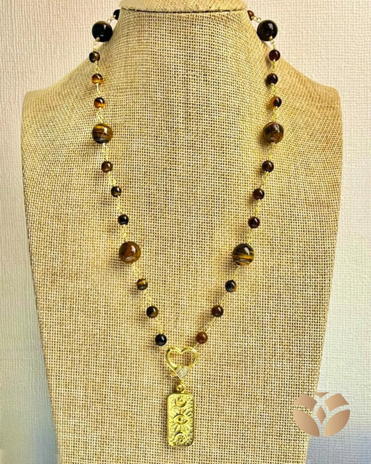 Está elaborado con piedras naturales Ojo de Tigre , cuidadosamente entorchadas para resaltar sus tonos dorados y marrones únicos.
En el centro, un broche en forma de corazón sostiene un dije dorado grabado con símbolos , que aporta un aire cómodo y distintivo.