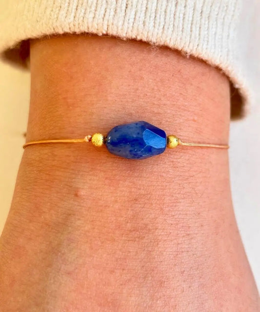 Pulsera de un hilo color beige que en el centro lleva una piedra de lapislazuli facetada y dos balies dorados a cada lado, posee cierre ajustable.