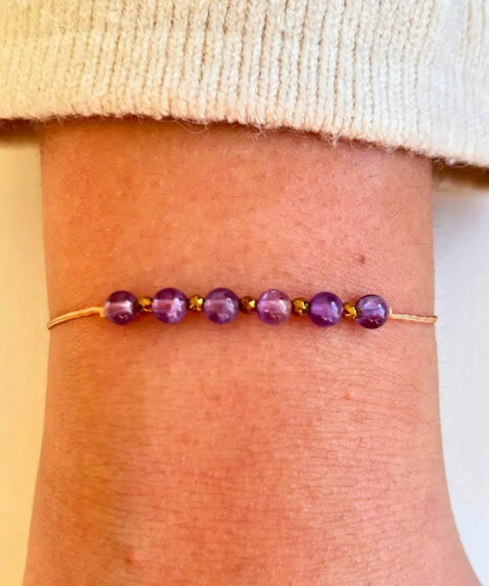 Pulsera de un solo hilo beige, lleva 6 piedras amatistas en el centro separadas por pequeños balines facetados en dorado profundo.